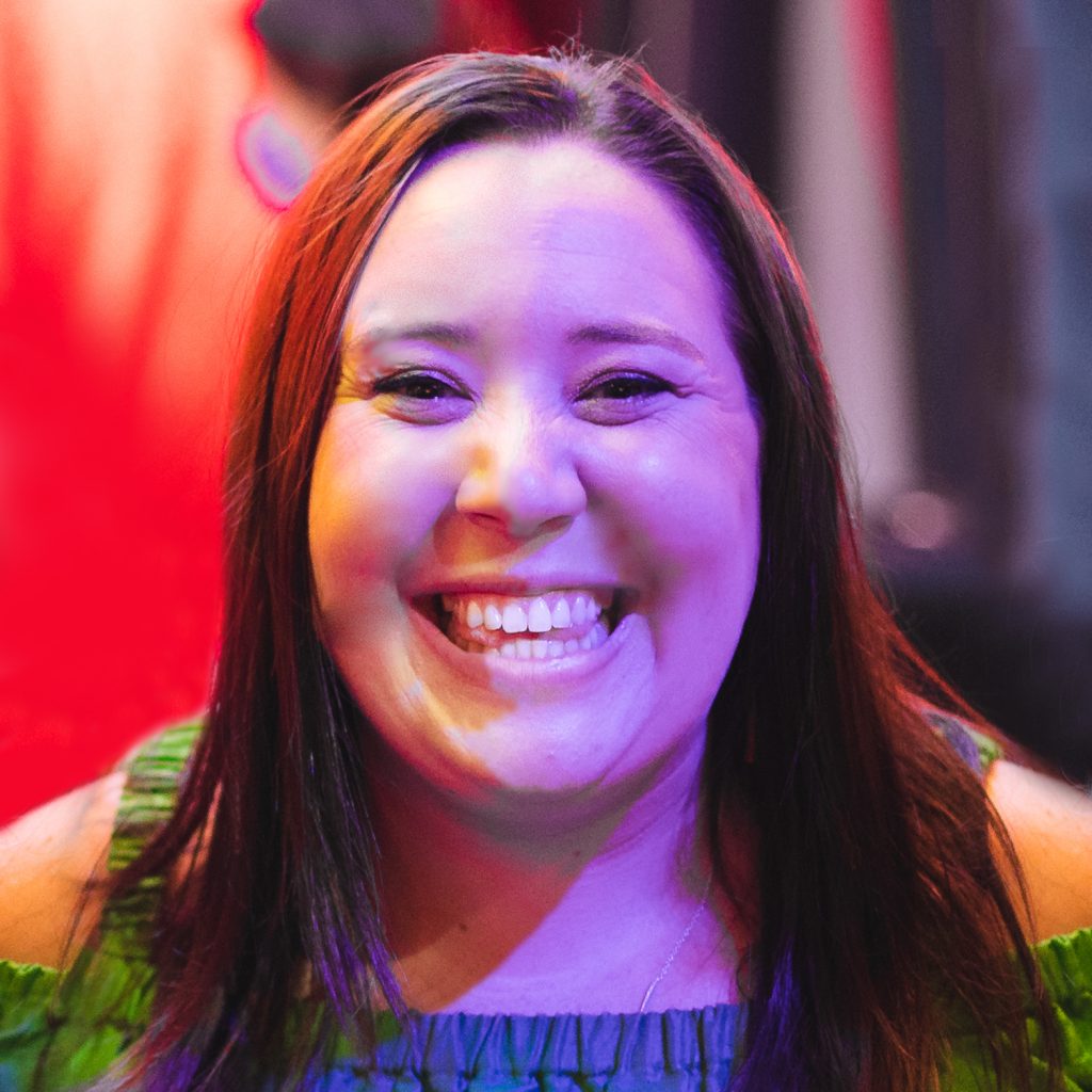 Smiling woman in colorful lighting, wearing a green top, with a blurred vibrant red background.