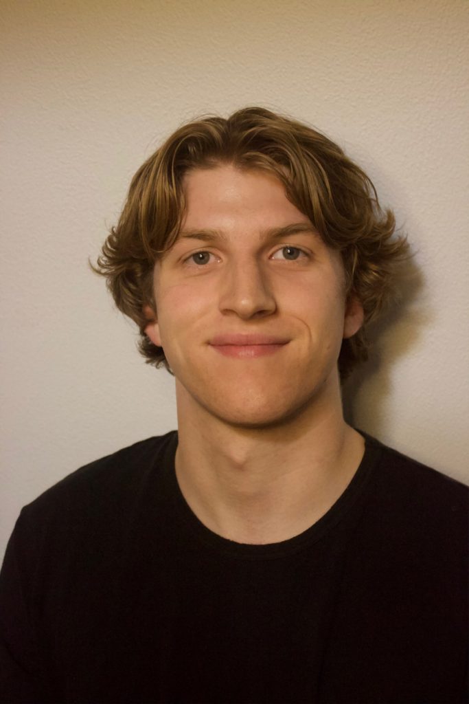 Smiling young person with medium-length hair and black shirt against a plain background.